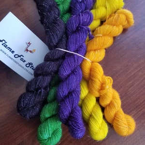 Puede incluir: Cuatro ovillos de hilo brillante en negro, verde, morado y amarillo. El hilo está dispuesto en una pila, con los extremos de los ovillos superpuestos. El hilo está etiquetado con "Flame Fox Studio" en una tarjeta blanca.