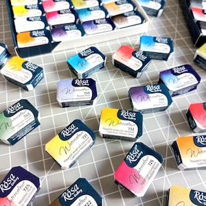 May include: A collection of watercolour paint pans in various colours, including yellow, orange, red, purple, blue, and green. The pans are labelled with the brand name "R&oacute;sa Gallery" and the colour name and number. The pans are arranged on a grey surface.