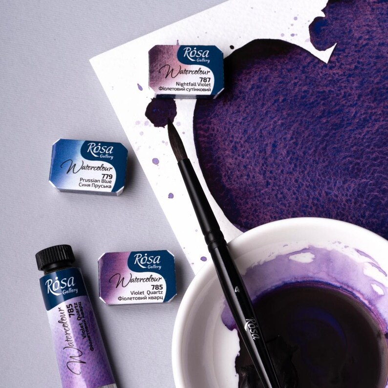 May include: Four watercolor paint tubes and pans in various shades of purple and blue. The tubes and pans are labeled with the brand name "Rosa Gallery" and the color name and number. The paint is in a white ceramic dish and a brush is on a white surface.