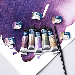May include: Six tubes of Rosa Gallery watercolor paint in various colors, including light violet, green quartz, cobalt brown, carbon yellow, and two shades of purple. The tubes are arranged in a row on a white surface with a purple watercolor wash in the background. A black paintbrush is also included.
