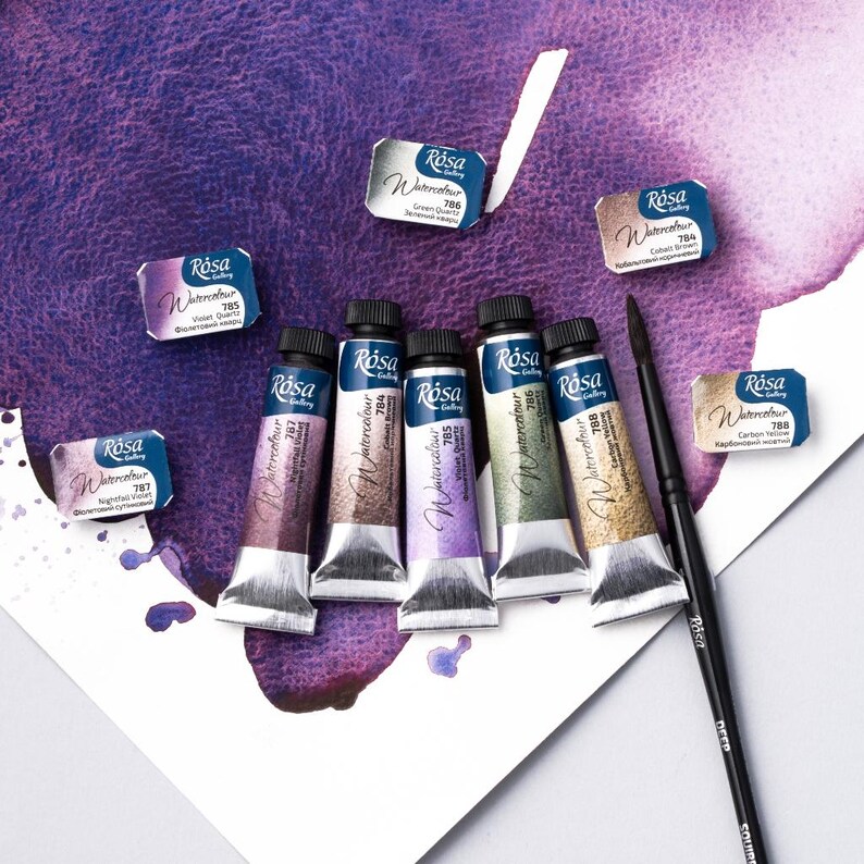 May include: Six tubes of Rosa Gallery watercolor paint in various colors, including light violet, green quartz, cobalt brown, carbon yellow, and two shades of purple. The tubes are arranged in a row on a white surface with a purple watercolor wash in the background. A black paintbrush is also included.