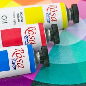 May include: Three tubes of Rosa Studio oil paint in yellow light, light red, and light blue, each with a black screw cap, are arranged diagonally across a circular color wheel featuring various painted segments in shades of blue, green, and purple.