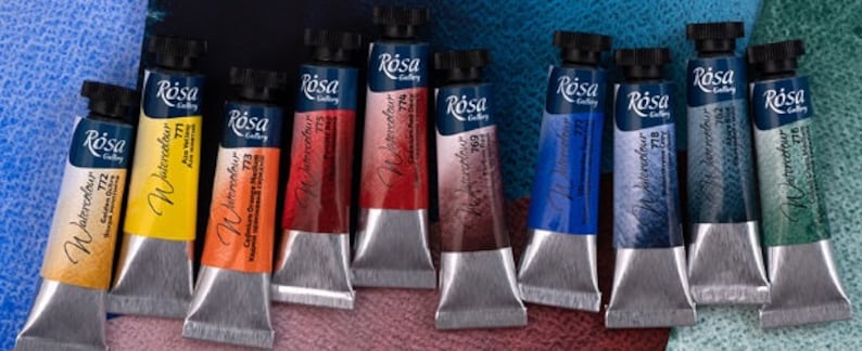 May include: Ten tubes of watercolor paint in various colors, labeled "R&oacute;sa Gallery Watercolor" with different numbers. The tubes are arranged in a row on a textured surface.