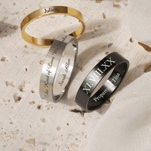 May include: Four engraved rings in gold, silver, black, and rose gold. The rings have personalized text, including names and phrases. The rings are displayed on a textured surface and white fabric.