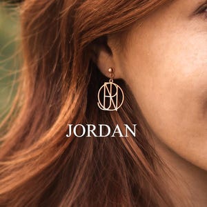 May include: A gold-colored earring with a circular design and the name "JORDAN" printed below it. The earring is worn by a person with red hair and fair skin. The background is blurred, with green and brown tones.