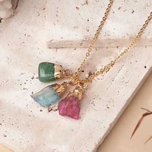 May include: A gold chain necklace with three gemstone pendants. The pendants are a green, blue, and pink stone.