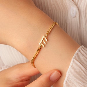 May include: Gold-colored chain bracelet with the number 777 as a charm. The bracelet is worn on a wrist, and the chain has a curb link design. The background is a white blouse.