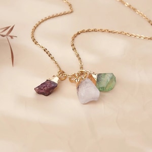 May include: A gold chain necklace with three different colored crystal pendants. The crystals are a deep red, a white, and a green.