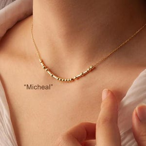 May include: A delicate gold necklace with a Morse code design, spelling "Micheal". The necklace features small, evenly spaced beads and cylindrical bars, strung on a fine chain. The necklace is worn around the neck.