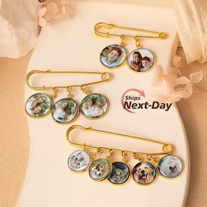 May include: Three gold-toned safety pins, each featuring circular charms with printed photographs. The top pin has two charms, the middle pin has three, and the bottom pin has four. The charms display various portraits of people and pets.