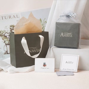 May include: A green gift bag with white handles and the word "ANAVIA" printed in gold. The bag is filled with brown tissue paper. A small white card with the words "A gift for you" is in front of the bag. A gray box with the word "ANAVIA" printed in white is in the background. A white box with the word "ANAVIA" printed in black is in the foreground. The box has the words "Jewelry Polishing Cloth" printed below the logo.