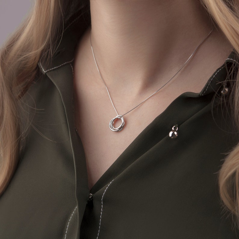May include: A silver necklace with a pendant featuring three interlocking rings. The necklace is worn by a person with long blonde hair and a green shirt.
