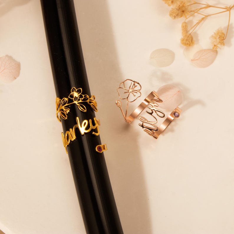 Puede incluir: Dos anillos de oro con delicados dise&ntilde;os florales y los nombres "Harley" y "Alyssa" inscritos en ellos. Cada anillo presenta una peque&ntilde;a piedra preciosa p&uacute;rpura.