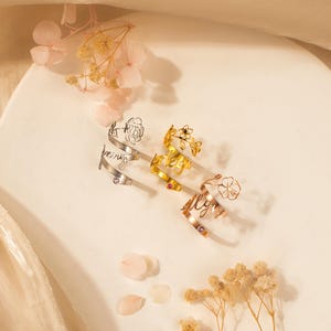 Puede incluir: Tres anillos de oro con delicados dise&ntilde;os florales y las palabras "always", "forever" y "being" inscritas en ellos. Cada anillo presenta una peque&ntilde;a piedra preciosa rosa.