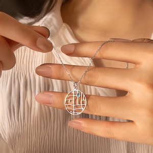 May include: A silver necklace with a geometric pendant. The pendant is a circle with a grid-like design and a small, round, blue gemstone. The necklace is held by a person wearing a white shirt.