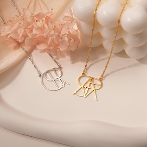 May include: Two gold necklaces with a delicate chain and a monogram pendant. The pendant is a stylized design of two letters intertwined. One necklace has a silver pendant and the other has a gold pendant.