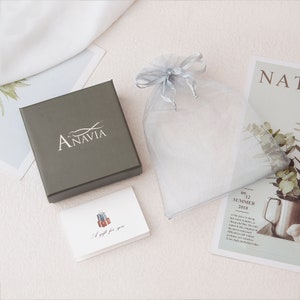 May include: A gray gift box with the word "ANAVIA" printed on the lid. The box is sitting on a white surface next to a small white card with the words "A gift for you" and a drawing of a suitcase. A gray organza bag is also visible in the image.