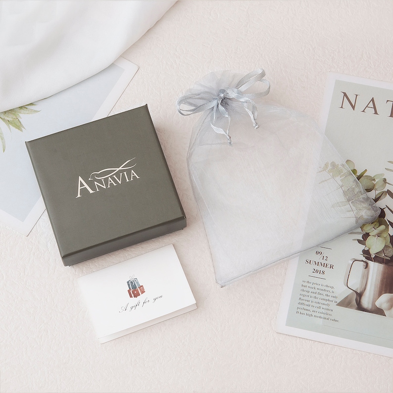 May include: A gray gift box with the word "ANAVIA" printed on the lid. The box is sitting on a white surface next to a small white card with the words "A gift for you" and a drawing of a suitcase. A gray organza bag is also visible in the image.