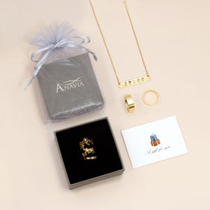 Puede incluir: Un conjunto de joyas de oro en una caja gris con una tarjeta de regalo blanca que dice "Un regalo para ti". Las joyas incluyen un collar con un colgante de barra, un anillo y un anillo en una caja. La caja tiene la palabra "Anavia" impresa en ella.