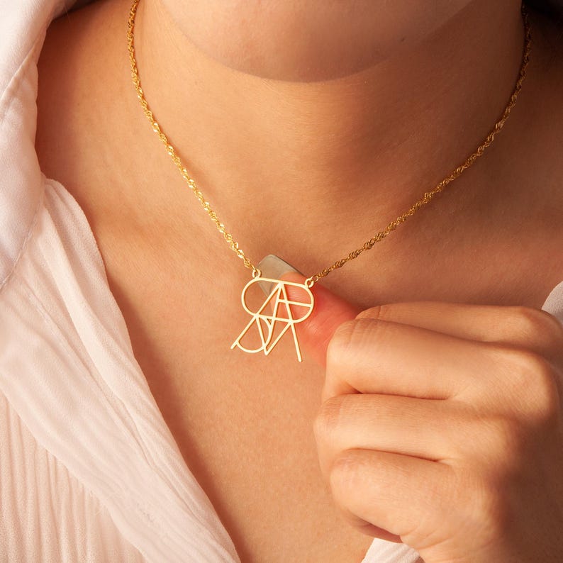 May include: A gold necklace with a geometric pendant featuring the letters "SA" intertwined. The necklace is on a person's neck.