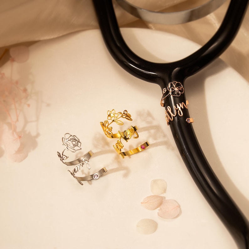 Puede incluir: Tres abalorios de estetoscopio dorados con dise&ntilde;os florales y nombres personalizados. Los abalorios son "Ashley", "Amber" y "Brittany".