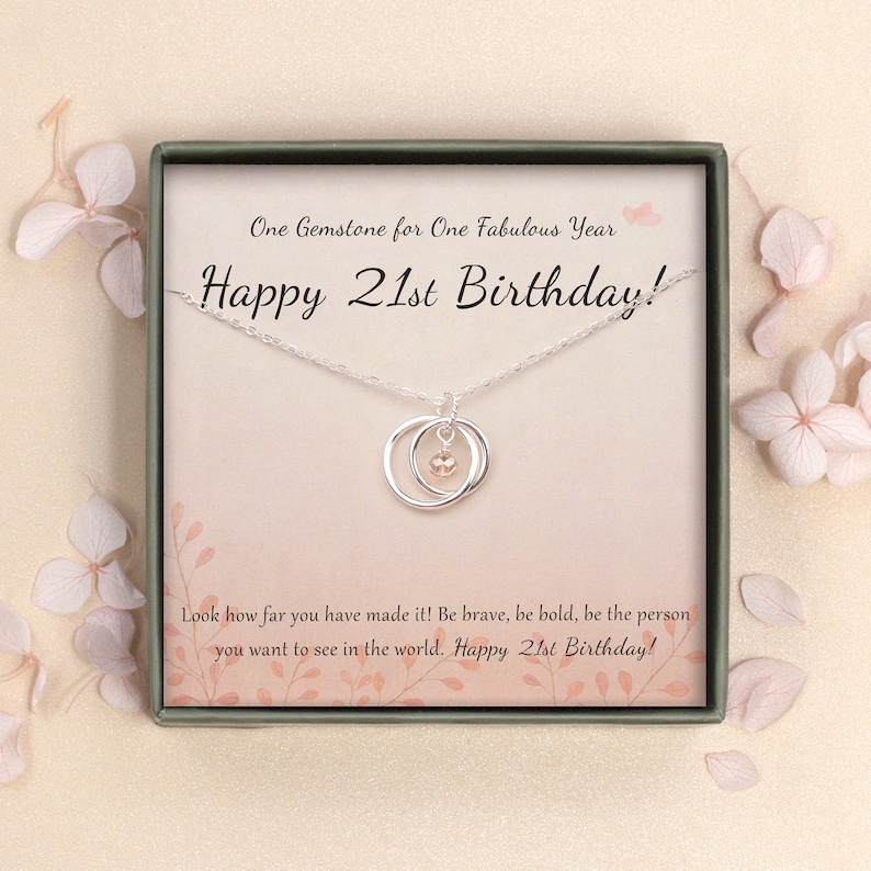 May include: A silver necklace with a double circle pendant and a pink gemstone. The necklace is in a white box with a green border and the text "One Gemstone for One Fabulous Year Happy 21st Birthday!" and "Look how far you have made it! Be brave, be bold, be the person you want to see in the world. Happy 21st Birthday!"