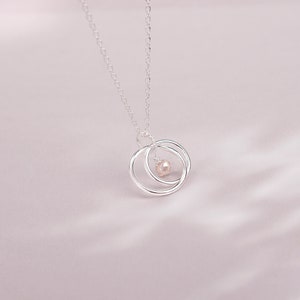 May include: A silver necklace with a delicate chain and a pendant featuring two interlocking circles with a small pearl in the center.