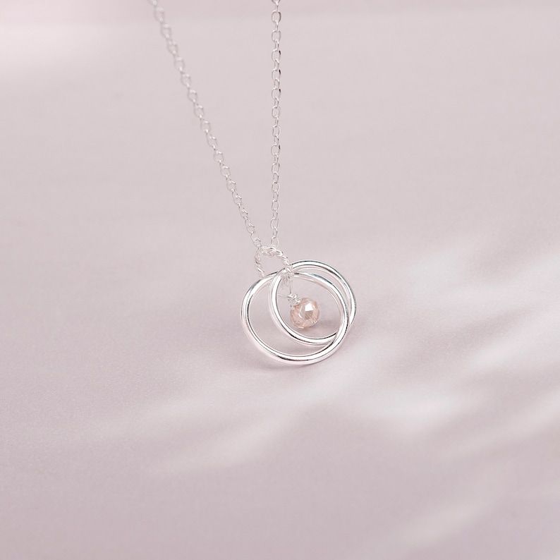 May include: A silver necklace with a delicate chain and a pendant featuring two interlocking circles with a small pearl in the center.