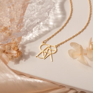 May include: A gold necklace with a geometric design pendant featuring the letters "RA". The necklace is on a white surface with a light brown fabric in the background.