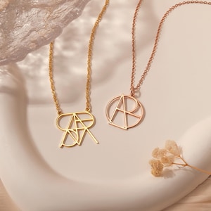 May include: Two gold necklaces with geometric letter pendants. One necklace features a single letter "A" pendant, while the other necklace features a double letter "AR" pendant.