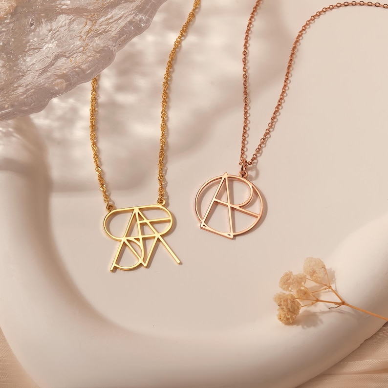 May include: Two gold necklaces with geometric letter pendants. One necklace features a single letter "A" pendant, while the other necklace features a double letter "AR" pendant.