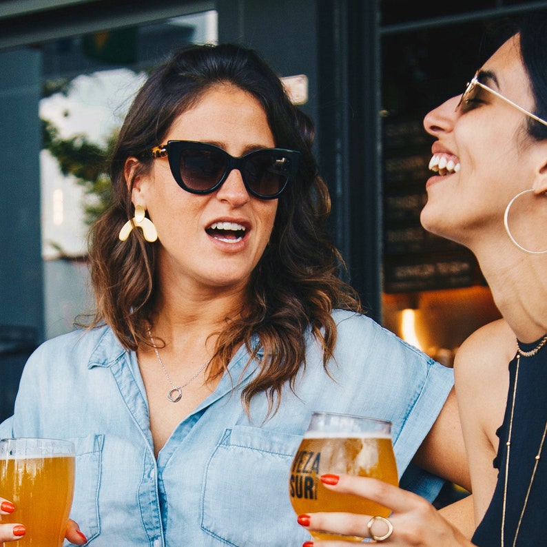 May include: Two women laughing and talking while holding glasses of beer. One woman is wearing black sunglasses and gold earrings. The other woman is wearing a black tank top and has a gold necklace. The beer glass in the foreground has a black and white label that says "Cerveza Sur".