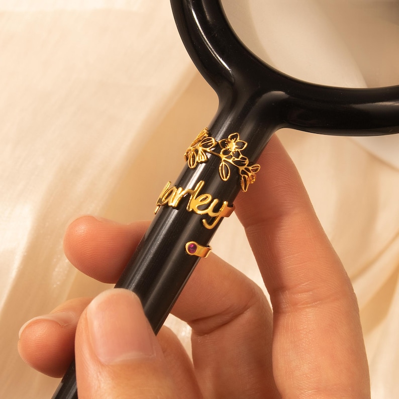 Puede incluir: Anillos dorados con un dise&ntilde;o floral y el nombre "Harley" en un estetoscopio negro.