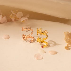 Puede incluir: Dos anillos de oro con delicados dise&ntilde;os florales y los nombres "Lily" y "Lexi" inscritos en ellos. Cada anillo tiene una peque&ntilde;a piedra preciosa rosa.