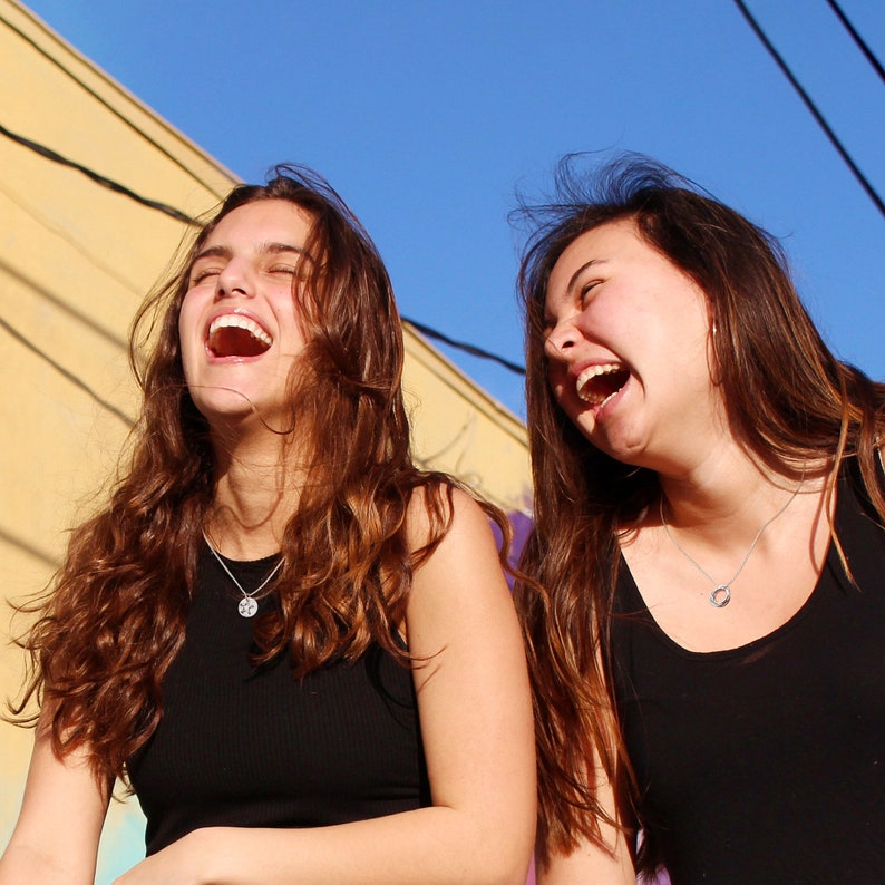 May include: Two women with long brown hair are laughing with their mouths open. They are wearing black tank tops and are standing outside against a blue sky.