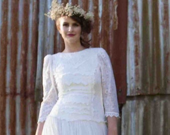 May include: A white lace wedding dress with a long skirt and a fitted bodice. The dress has a scalloped neckline and long sleeves with lace trim. The bride is wearing a flower crown.