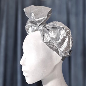 May include: A silver headband with a large bow. The headband is made of a shiny, textured fabric.