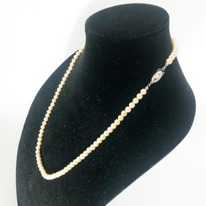 May include: A delicate pearl necklace displayed on a black velvet bust. The necklace features numerous small, creamy-colored pearls strung together. The clasp is silver-toned with a decorative design. The background is plain white.