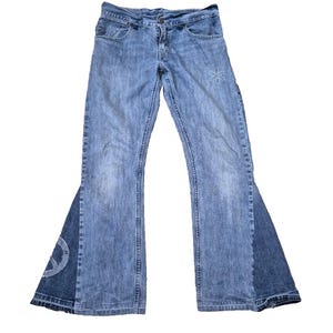 Op de afbeelding: Een paar lichtblauwe denim bell-bottom jeans met een witte geborduurde bloem op het rechterbeen en een wit geborduurd vredesteken op het linkerbeen.