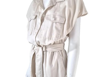 May include: A light beige jumpsuit with short sleeves, a collared neckline, and two chest pockets. The jumpsuit features a button-down front, a belted waist, and two front pockets. The fabric appears to be a lightweight cotton or linen blend.