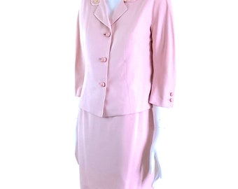May include: A pale pink two-piece suit. The jacket features a notched collar with floral embroidery, three buttons, and three-quarter sleeves. The matching skirt is knee-length. The suit is displayed on a mannequin.