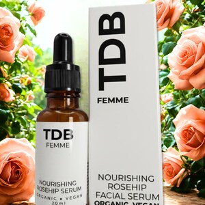May include: A brown glass bottle with a black dropper and a white label, next to a white box. Both feature the text "TDB FEMME" and product details. The bottle contains 20 ml of Nourishing Rosehip Serum, and the box describes it as an Organic Vegan Facial Serum.