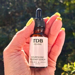 May include: A brown glass bottle of 'TDB Femme' Nourishing Rosehip Serum, 20ml, held in a hand with yellow nail polish. The bottle has a black dropper and a white label with black text. The background is blurred green foliage.