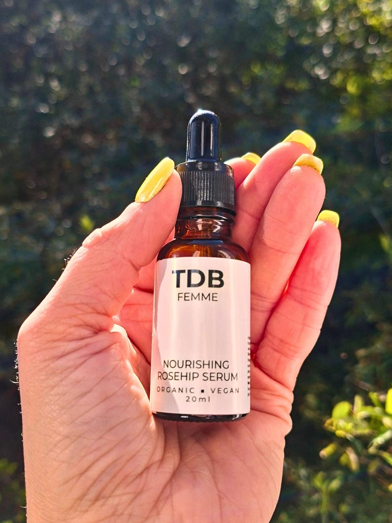 May include: A brown glass bottle of 'TDB Femme' Nourishing Rosehip Serum, 20ml, held in a hand with yellow nail polish. The bottle has a black dropper and a white label with black text. The background is blurred green foliage.