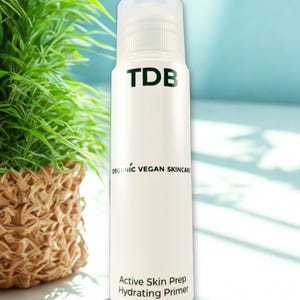 Puede incluir: Botella cilíndrica blanca de producto para el cuidado de la piel con tapón blanco. La botella tiene el texto "TDB" en negro, y "ORGANIC VEGAN SKINCARE" y "Active Skin Prep Hydrating Primer" en negro. El fondo es azul claro.