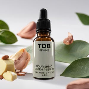 May include: A brown glass bottle of TDB Femme Nourishing Rosehip Serum with a black dropper. The label reads "Organic Vegan 20ml". The background includes green leaves and nuts.