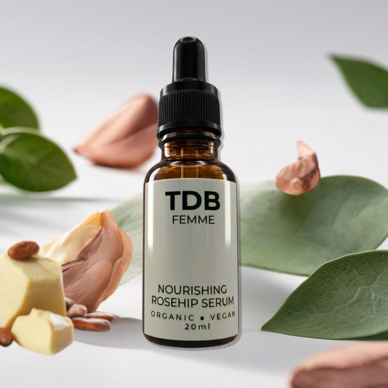 May include: A brown glass bottle of TDB Femme Nourishing Rosehip Serum with a black dropper. The label reads "Organic Vegan 20ml". The background includes green leaves and nuts.