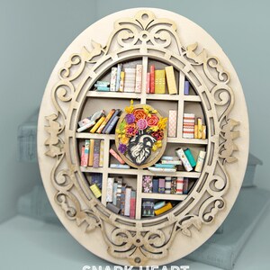 May include: A round wooden wall decor with a carved ornate frame. The center features a miniature bookshelf filled with colorful books and a floral arrangement with a heart in the center. The text "SNARK HEART" is printed below the frame.