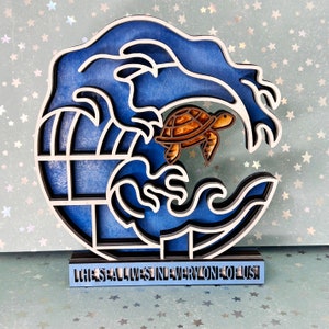 May include: A wooden wall art piece depicting a blue wave with a turtle inside. The wave is outlined in black and the turtle is brown. The base of the piece is blue and reads "The Sea Lives in Everyone of Us".