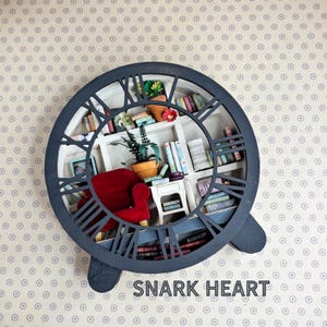 Peut inclure: Un cadran d'horloge noir avec des chiffres romains entoure une pièce miniature avec une bibliothèque, un fauteuil rouge, une plante en pot et une petite table. Le cadran de l'horloge est monté sur un support noir à deux pieds. Le texte "SNARK HEART" est imprimé sous l'horloge.